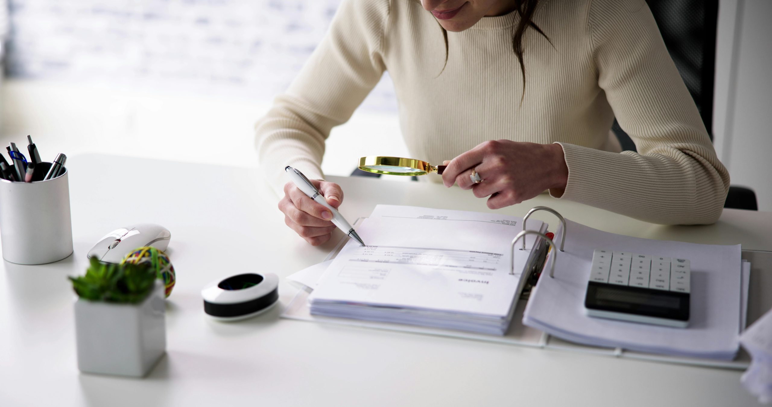 A woman in an office meticulously examines financial statements and legal documents with a magnifying glass, emphasizing detailed scrutiny of assets during a legal process.