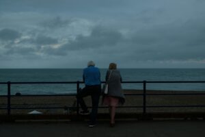 The backs of an elderly couple standing at a railing and looking out over a tumultuous sea in different directions under a dark and cloudy sky, representing grey divorce in Ontario.