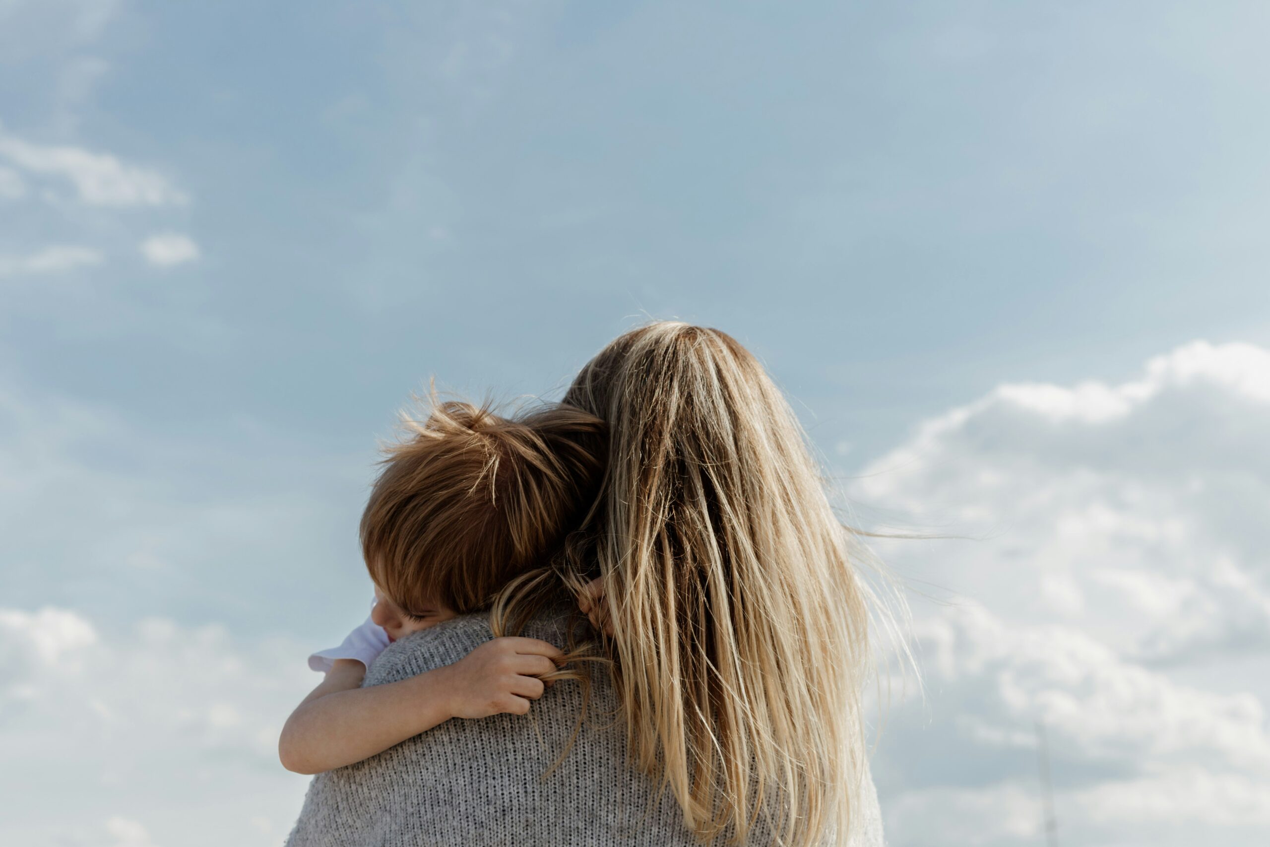 A woman with long blonde hair holding her young son in her arms, looking up into a wide blue sky, representing the support Johnson Miller Family Lawyers provides post-separation or divorce, including separation agreements that address parenting issues, property division, child support, and spousal support.