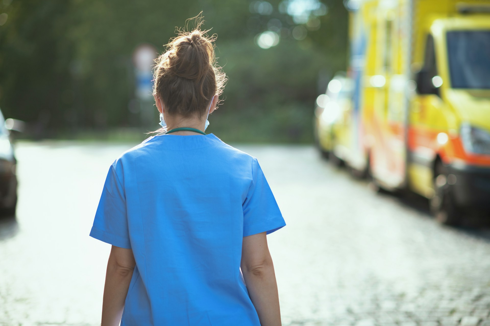 A nurse, from the back and wearing blue scrubs, walks past blurred emergency vehicles, representing parenting schedules for shift workers.
